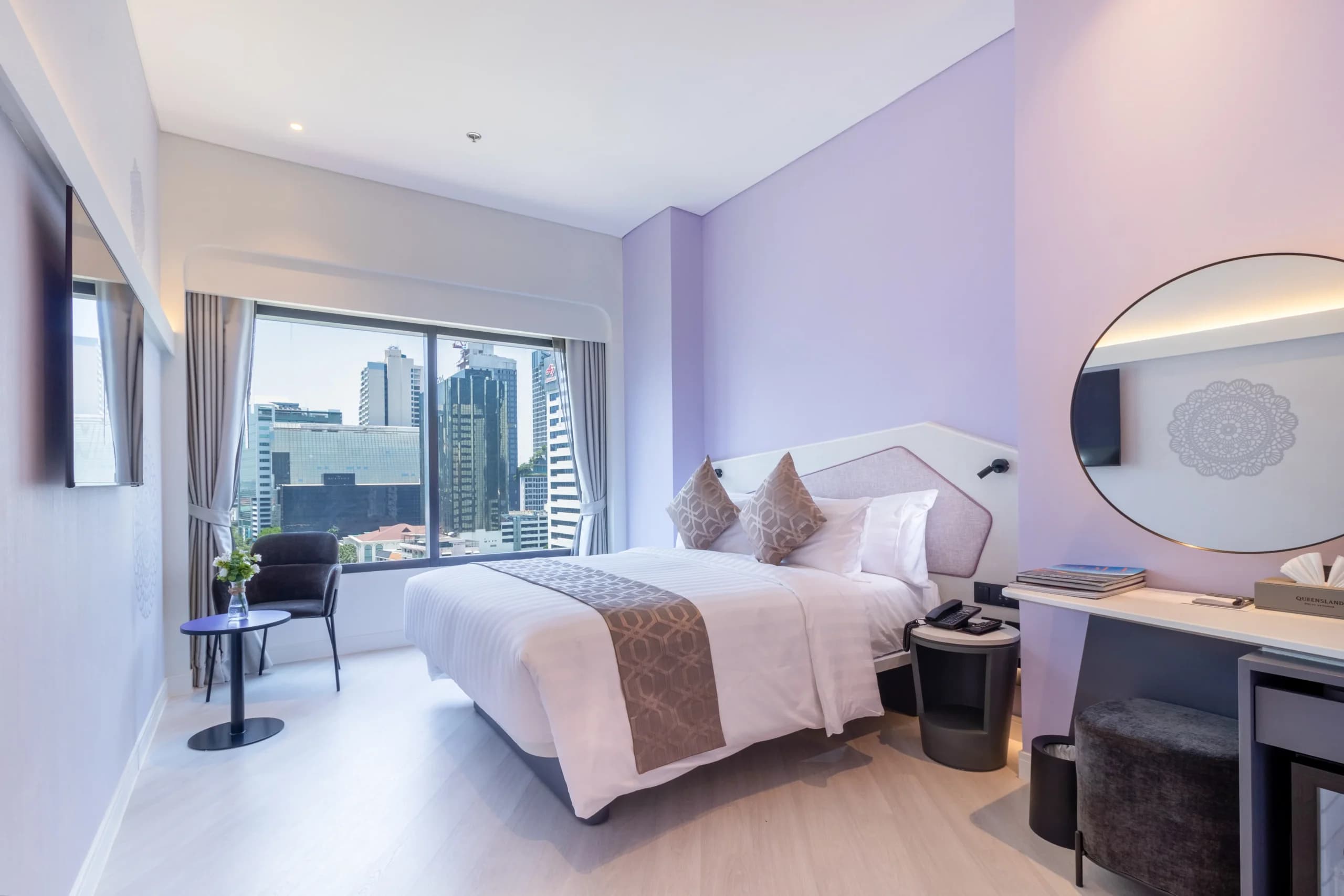 A guest room with a bed, a round mirror, a desk and a seat by the window.