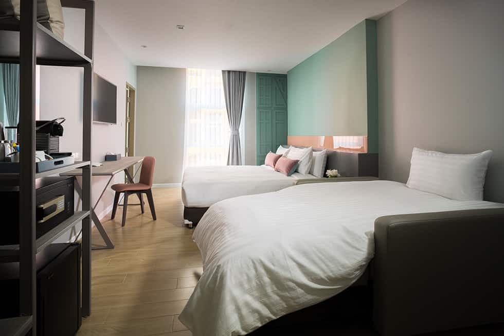 A room with a large bed, a sofa bed and a desk beside a bright window.